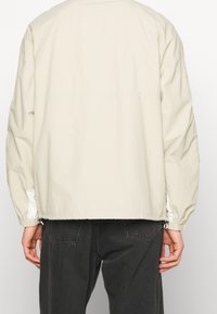 Persona vistiendo una chaqueta de manga larga en color beige claro y unos jeans de mezclilla negros, de pie de espaldas a la cámara contra un fondo plano.