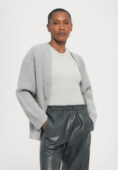 Hele hallikas soonikko cardigan, millel on ülekäte varrukad ja kaks nööpi, kantud sobiva helehall särk ja läikivad tumeda halli püksid.