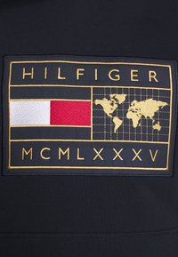 Tmavě modrá látka s vyšitým obdélníkovým logem zobrazujícím mapu světa, zlaté akcenty a červeno-bílé pruhy. Text zní "HILFIGER MCMLXXXV."