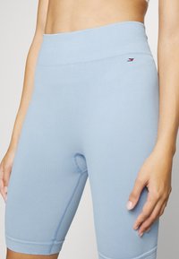 Femme portant un short de cycliste bleu clair côtelé à taille haute avec un petit logo sur la hanche gauche, main reposant sur le côté, cadrée à mi-taille et cuisses.
