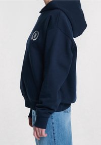 Sudadera con capucha de color azul marino y corte holgado, con un logo bordado y puños. Tela suave con textura lisa.