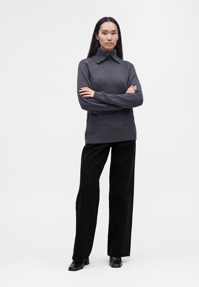 Femme debout, bras croisés, portant un pull gris foncé à large col, un pantalon noir à jambes larges et des chaussures noires sur un fond uni.