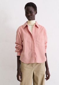 Light pink button-up shirt with a pointed collar, single chest pocket, and rolled-up sleeves, paired with beige wide-leg trousers.