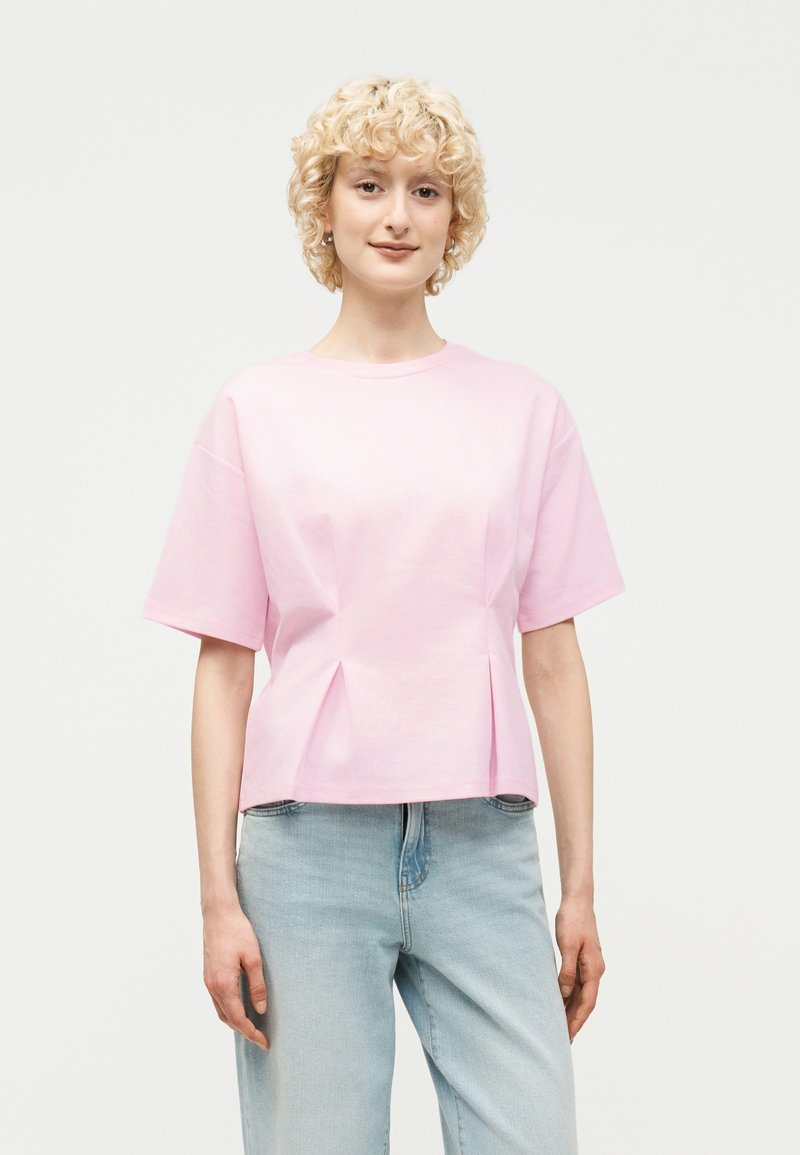 Νεαρό άτομο με κοντά σγουρά ξανθά μαλλιά, φορώντας ένα ανοιχτό ροζ κοντομάνικο πουκάμισο και ανοιχτό μπλε τζιν, στέκεται μπροστά σε απλό φόντο.