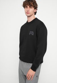 Μαύρο φούτερ με στρογγυλή λαιμόκοψη, υφασμάτινη υφή και κεντημένη λευκή λεπτομέρεια λογότυπου. Συνδυάζεται με ανοιχτό γκρι φόρμα με έμφαση σε λογότυπο.