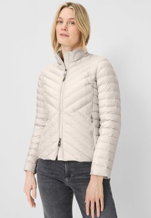 Woman wearing a light beige quilted jacket with a high collar, zipped up, paired with dark gray jeans, standing against a plain background.