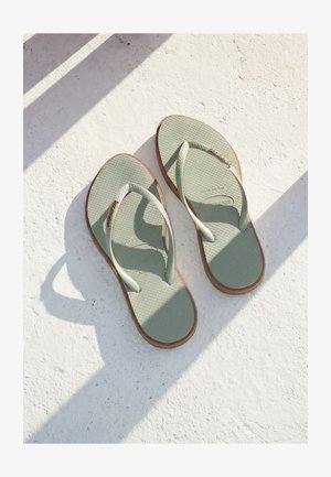 Paar hellgrüne Flip-Flops mit braunen Sohlen auf strukturierter weißer Oberfläche mit diagonalen Schatten.
