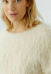 Nahaufnahme einer Frau mit blonden Haaren, die einen weichen, flauschigen cremefarbenen Pullover mit geripptem Halsausschnitt vor einem einfarbigen, hellen Hintergrund trägt.