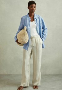 Camicia di lino azzurro chiaro, canotta bianca, pantaloni beige con coulisse, borsa in paglia intrecciata e sandali marroni, mostrati su uno sfondo neutro.