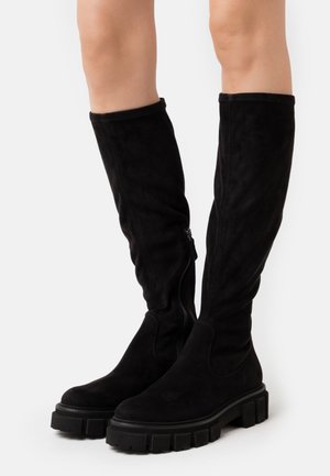 Nogi w czarnych zamszowych kozakach za kolano z masywną, protektorowaną podeszwą i zamkami bocznymi na białym tle.