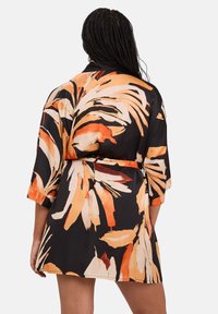 Personne avec des cheveux tressés portant une robe noire courte avec ceinture, ornée de grands motifs floraux abstraits orange et beige, vue de dos.