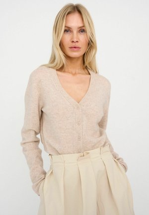 Béžový pletený kardigán s výstrihom do V, zapínaním na gombíky a rebrovanými manžetami. Kombinovaný s vysokými, plisovanými svetlo béžovými nohavicami.
