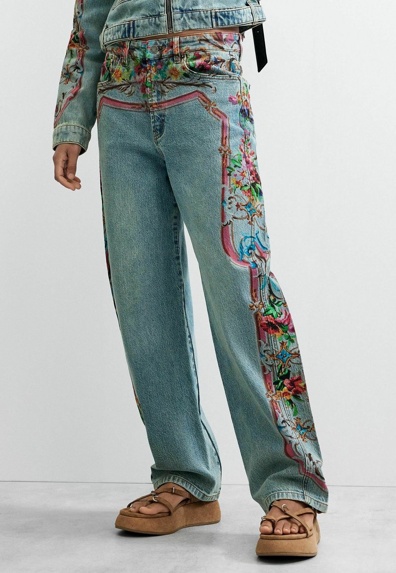 Denim jeans with colorful floral and ornamental patterns on sides and pockets, paired with brown platform sandals.
