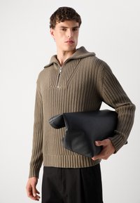 Bež pleteni pulover s ovratnikom na zadrgo, s rebrastom teksturom i dugim rukavima. Model drži crnu kožnu torbicu koja izgleda poput jastuka.