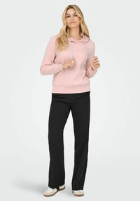 Lys rosa hettegenser med sølv snorer, featuring et hjerte-logo. Kombinert med svarte bukser og hvite joggesko med brune såler.