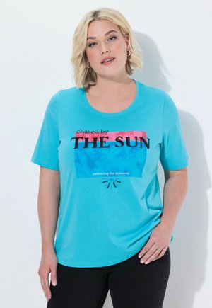 Lyshåret kvinde iført en knaldblå T-shirt med teksten "chased by THE SUN embracing the unknown" og sorte bukser, stående mod en hvid væg.