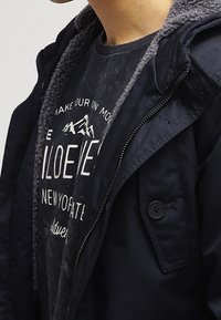 Giacca blu navy con fodera in pile grigio dalla texture. Presenta una chiusura con zip, tasche laterali e un dettaglio con bottone sul petto sinistro. Indossata sopra una t-shirt con stampa grafica.