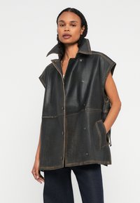 Donna in un gilet di pelle nera senza maniche con colletto alto, bottoni frontali, tasche e pantaloni larghi scuri, in piedi contro uno sfondo semplice.