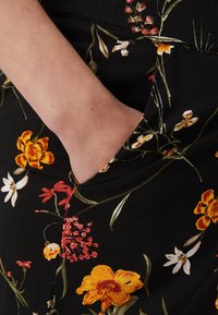Květinové šaty s černým pozadím a živými žlutými, červenými a bílými květinami. Obsahují boční kapsu a hladkou texturu tkaniny.