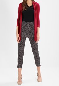 Červený otevřený cardigan, černý top s krajkovým lemem a černé kalhoty s potiskem. Kalhoty mají geometrický vzor. Oblečení doplňují světlé béžové podpatky.