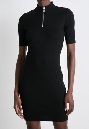 Vestido corto ajustado de manga corta en negro con cuello alto y cremallera frontal plateada, llevado por una persona de pie sobre un fondo liso.