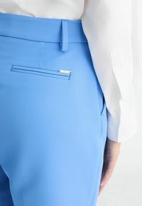Světle modré kalhoty na míru s nohavicemi obsahujícími zadní kapsu se stříbrným akcentem, vyrobené z hladké látky, v kombinaci s bílou blůzou.