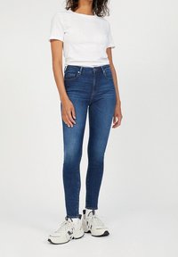 Úzké modré džíny, vysoký pas, mírně mechanicky opotřebovaný vzhled, kombinované s jednoduchým bílým tričkem a bílými teniskami.