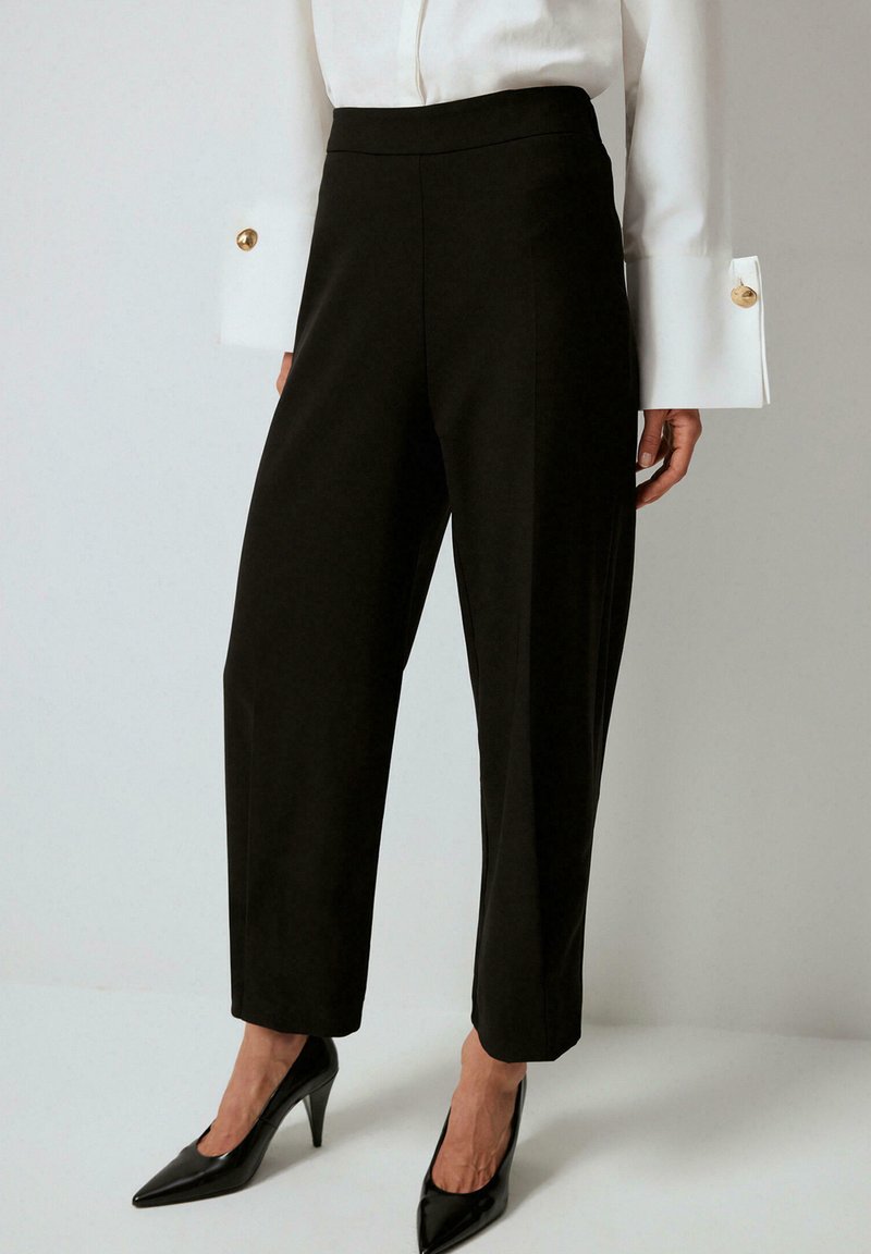 Black tapered trousers with a high waist, featuring a smooth fabric finish. Accented by a white blouse with large cuffs and gold buttons.