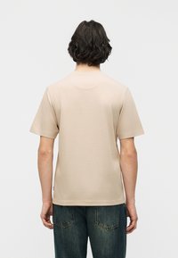 Άτομο με σκουρόχρωμα σγουρά μαλλιά στέκεται με την πλάτη γυρισμένη, φοράει απλό μπεζ κοντομάνικο πουκάμισο και σ κούρα μπλε τζιν, μπροστά σε λευκό φόντο.