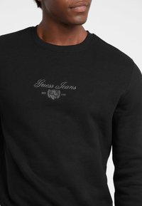 Felpa nera in cotone, con colletto rotondo, polsini a coste e logo "Guess Jeans" ricamato in grigio chiaro.