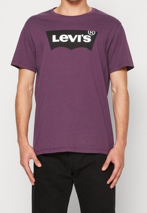 Mężczyzna w fioletowej koszulce Levi's z czarno-białym logo, ubrany w czarne spodnie, stoi na jednolitym białym tle.