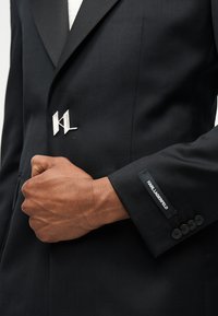 Χέρι άνδρα που προσαρμόζει μαύρο σακάκι Karl Lagerfeld με τέσσερα κουμπιά και ασημένιο σήμα KL στο πέτο.