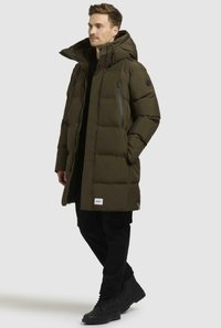 Giacca puffer verde oliva con colletto alto e chiusura frontale con zip. Presenta tasche sul petto con zip e una toppa con logo. Indossata con pantaloni neri e stivali.