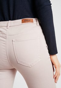 Jeans de mezclilla rosa claro con un bolsillo trasero y una etiqueta de cuero marrón etiquetada "ONLY". La tela tiene una textura suave y un corte regular.