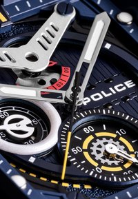 Analoge horloge met een donkerblauwe wijzerplaat, zilveren accenten, witte en gele uurmarkeringen en meerdere sub-wijzers. Bevat een prominent 'POLICE' logo.