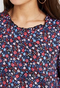 Femme portant un chemisier floral foncé avec des fleurs rouges, bleues et roses, un col festonné noir laissant entrevoir une partie du cou et une boucle d'oreille en créole.