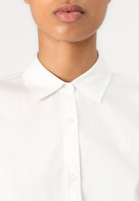 Gros plan d'une personne portant une chemise blanche à col boutonnée jusqu'en haut avec un logo subtil Hugo sur la poitrine.
