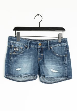 Short en jean bleu avec détails déchirés, fermeture à boutons, poches avant, suspendu à un cintre noir sur fond blanc.