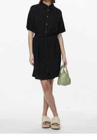 Abito nero a maniche corte con chiusura a bottoni, cintura in tessuto in vita e texture leggera. Il modello tiene una borsa in rete verde e indossa sandali con plateau beige.