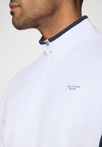 Fehér zipzáras dzseki sötétkék gallérral. Jellemzője egy kis márkás címke, amelyen az áll: "TED BAKER SPORT." Sima textúrájú és sportos dizájnú.