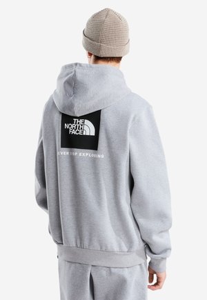 Osoba ubrana w szarą bluzę z kapturem The North Face i pasujące spodnie, zwrócona tyłem, na głowie beżowa dziergana czapka, widoczne logo i napis na plecach.