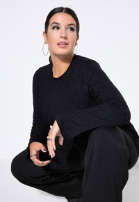 Schwarzes Langarmshirt mit strukturiertem Blumenmuster, kombiniert mit lockeren schwarzen Hosen. Das Model hat dunkles Haar, das zurückfrisiert ist, und trägt Creolen.