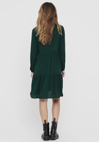 Robe verte à volant avec de longues manches et un ourlet festonné. Assortie à des bottines noires avec des étiquettes jaunes. Texture de tissu douce.