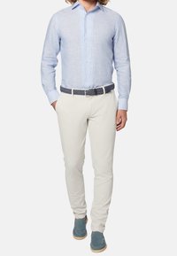 Mężczyzna w jasnoniebieskiej koszuli w paski z guzikami, białych spodniach z plecionym ciemnym paskiem i granatowych zamszowych loafersach, stojący z rękami w kieszeniach.