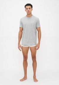 Graues Kurzarm-T-Shirt aus weichem Stoff, runder Halsauschnitt, kombiniert mit weißen Shorts, das Modell steht barfuß vor einem neutralen Hintergrund.