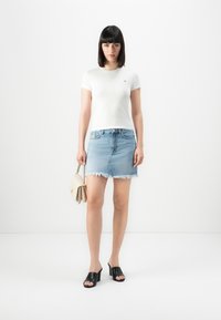 Femeie în picioare, purtând un tricou alb ajustat, o fustă din denim decolorat, de culoare albastru deschis, mule negre cu toc, ținând o geantă bej, pe un fundal alb.