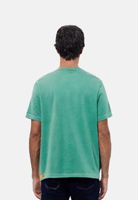 T-shirt in cotone verde con maniche corte e collo rotondo. Presenta una vestibilità rilassata e un'etichetta con il marchio sul lato sinistro inferiore.
