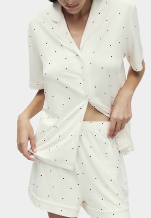 Vrouw die een crèmekleurige blouse met korte mouwen en bijpassende shorts met elastische taille draagt, beide met kleine zwarte polkadotjes.