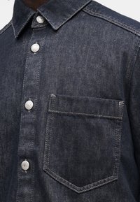 Tmavě modrá denimová košile s límečkem na knoflíky, stříbrnými patentkami a náprsní kapsou s kontrastním prošíváním.