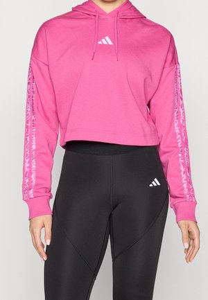 Kobieta w różowej krótkiej bluzie z kapturem z wzorzystymi paskami na rękawach oraz czarnych legginsach z wysokim stanem, oba z białymi logo Adidas.